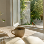Kaffeetasse und offenes Buch im warmen Morgenlicht, während ein Rotkehlchen am Fenster sitzt